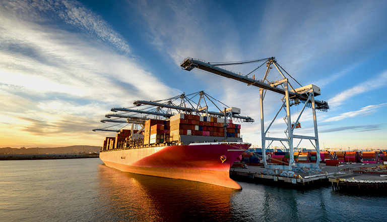 Aerial shot of a massive cargo ship loaded with containers docked in the Port of Long Beach, with the booms of ship-to-shore gantry cranes reaching out from the pier of the terminal