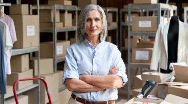 A woman smiling with her arms crossed in a warehouse with boxes on shelves.