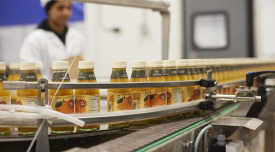 Glass bottles of organic orange juice on a machine in a factory setting with a blurred worker in the background.
