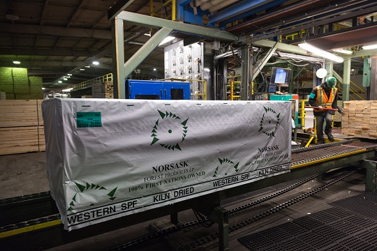 A pallet of lumber from Norsask Forest Products sits on a conveyer belt, with a worker in the background.