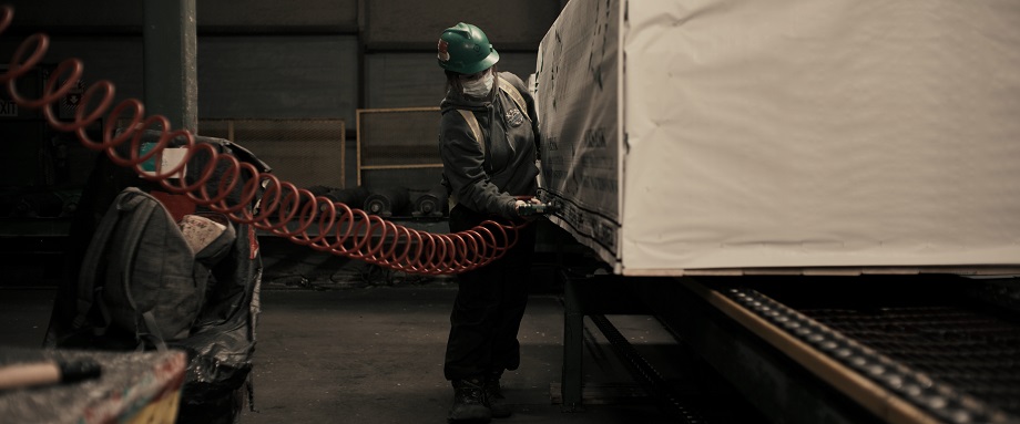A woman in a green hardhat and grey sweatshirt uses a compression tool with a red power cord to attach the paper cover to a shipping pallet of lumber.