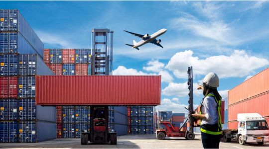 An airplane flying above shipping containers, trucks, and a worker wearing a safety vest on the ground.
