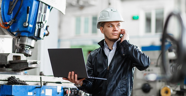 An engineer standing in front of an industrial machine with his laptop talks on the phone