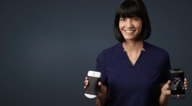 A smiling woman holding a handheld ultrasound scanner in one hand and a phone displaying a fetal ultrasound in the other.