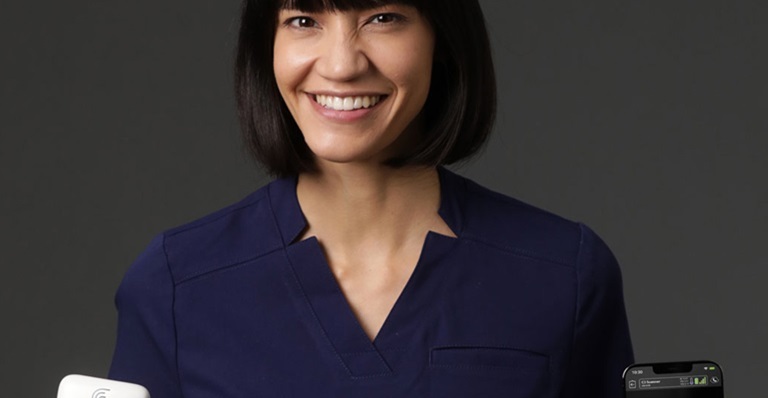 Smiling women holding Clarius portable ultrasound device in left hand and a smartphone in the right hand which shows an image of a fetal ultrasound.