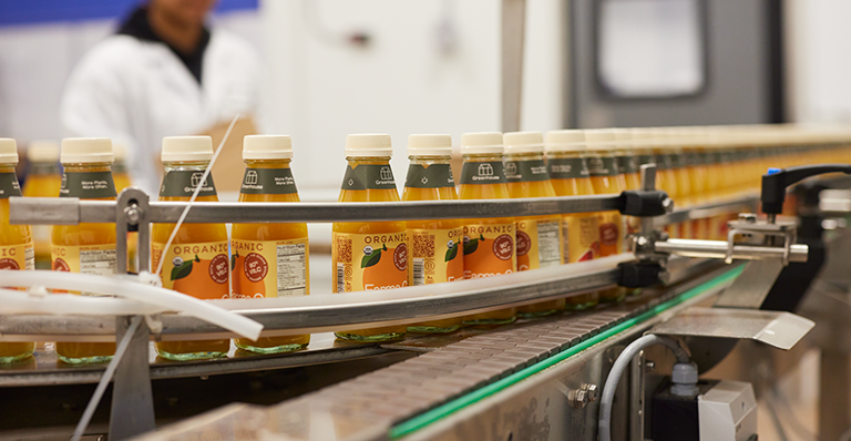 Bottles of organic orange juice on a conveyer belt. A woman in a white coat is in the background.  