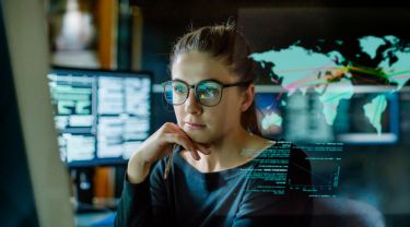 A young woman wearing glasses sitting in a dark office surrounded by computer monitors. In front of her is a transparent display showing a world map with overlaid data.