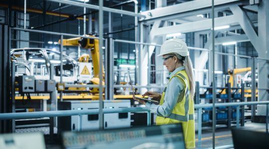 A woman wearing a hard hat, safety glasses, and a safety vest using a laptop, with an automated robot arm manufacturing a vehicle blurred in the background. 