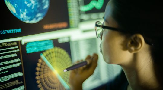 A woman wearing glasses, holding a pen and looking at data on a screen.