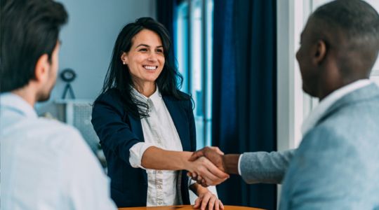 A businesswoman and a businessman shaking hands in an office, with another businessman standing nearby, observing the interaction.