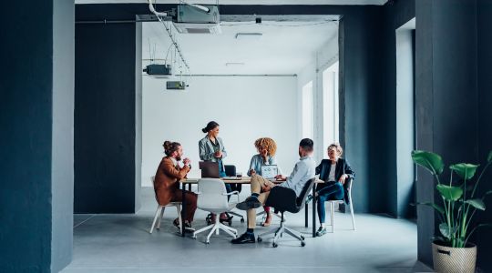 Un groupe de cinq professionnels assis et debout autour d’une table sur leurs ordinateurs portables.
