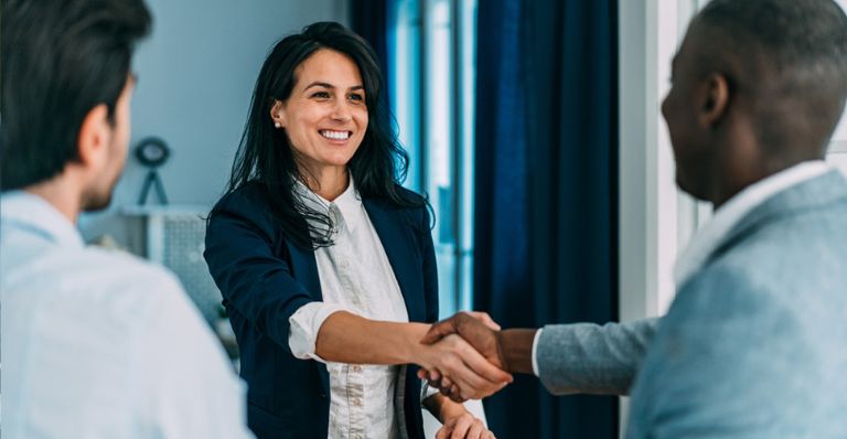 A businesswoman and a businessman shaking hands in an office, with another businessman standing nearby, observing the interaction.