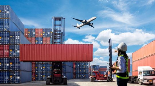 Un avion vole au-dessus de conteneurs et de camions, et une personne portant un gilet de sécurité se trouve au sol.