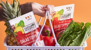A hand holding a white grocery basket filled with fruit, vegetables and naturSource products against an orange background.
