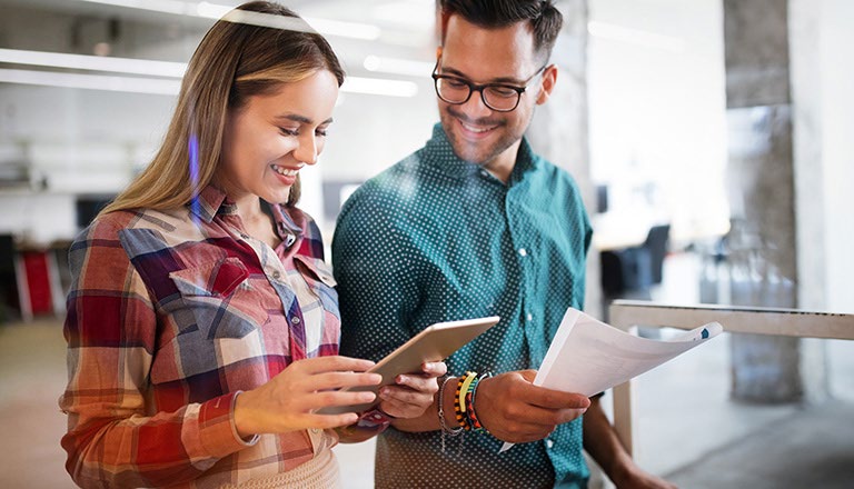 Two people smiling and looking at a tablet. One individual is holding a piece of paper.
