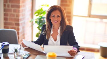 A woman smiling and sitting on a chair while reading documents at her desk.
