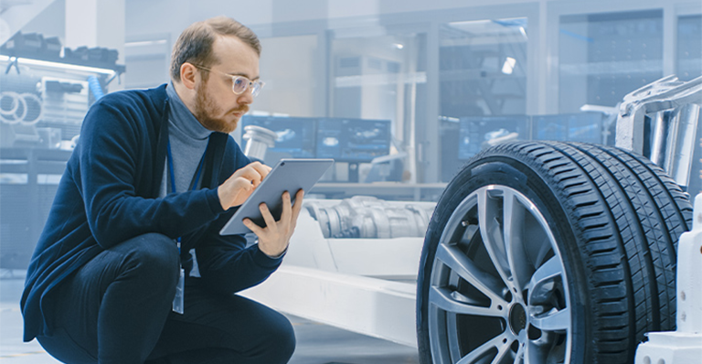 Man holding tablet inspects an automobile frame and wheel assembly in a factory.
