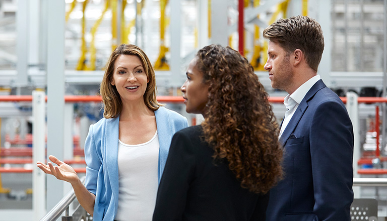 Mature businesswoman talking with coworkers at automobile industry. A smiling female entrepreneur is standing with colleagues by a railing. They are wearing business wear at a factory.