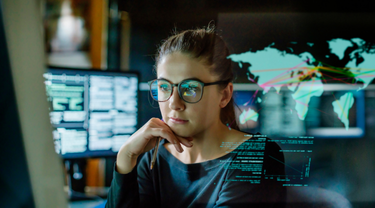 Young woman, wearing glasses, surrounded by computer monitors in dark office, with display showing world map.