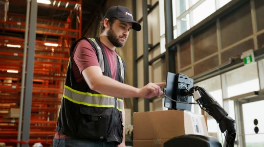 Man in a baseball cap and safety vest using a tablet in an inventory setting. 