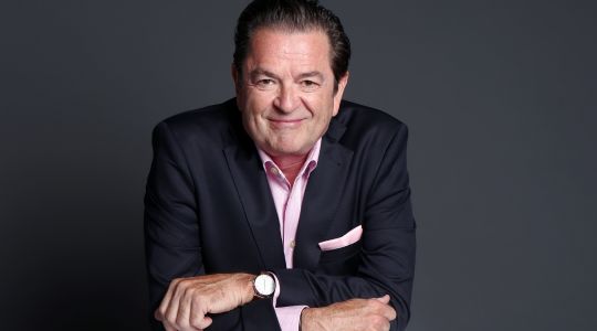 Un homme assis avec les bras croisés à un bureau, portant un costume, souriant à la caméra. 